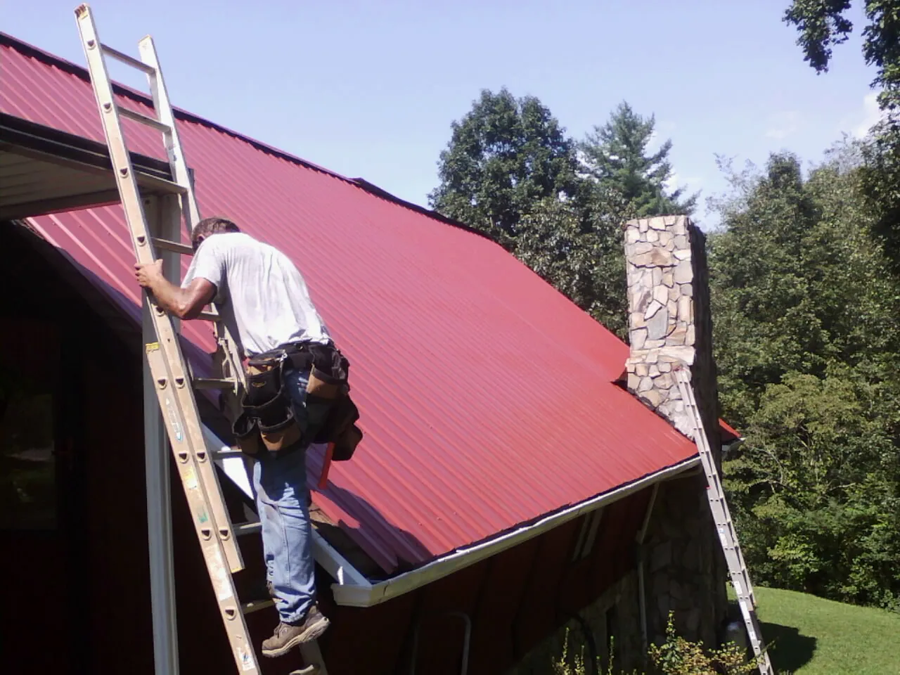 Roofer on a ladder beside a red metal roof and stone chimney — Soffit & Fascia Repair by Roofing Brothers in Missoula
